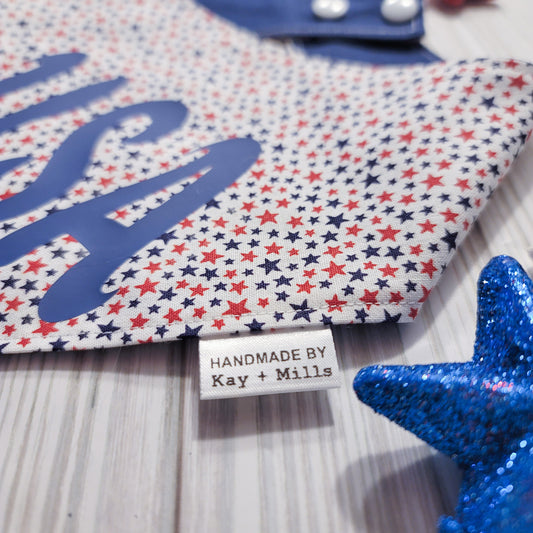 A close up image of pet bandana with small red and navy blue stars on a white background and USA written in vinyl. The back side of the bandana is navy blue and it has two sets of white plastic snaps. A brand tag is attached to the side of the bandana stating "Handmade by kay + mills".