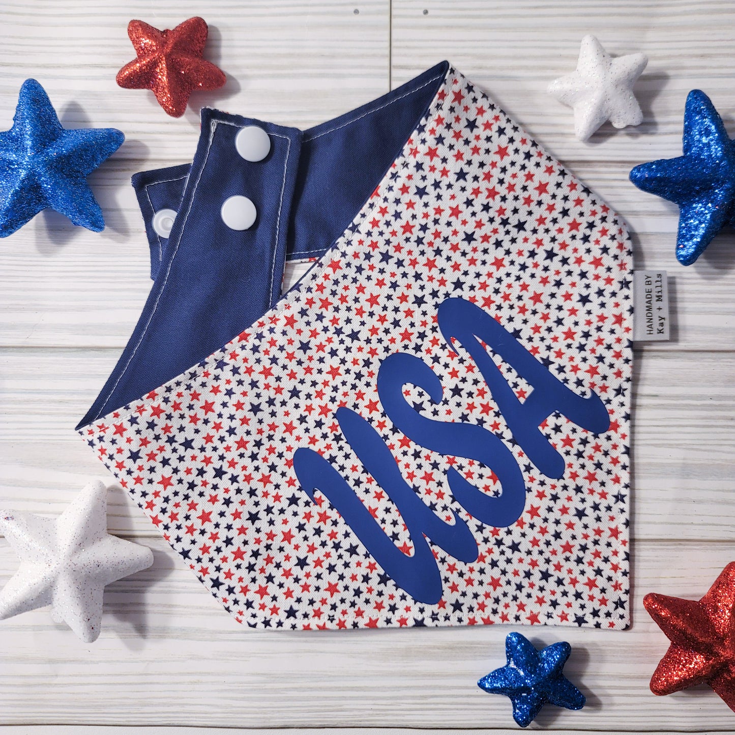 Dog bandana or cat bandana with small red and navy blue stars on a white background and the letters USA in navy blue vinyl. The back of the pet bandana is navy blue and includes two sets of white plastic snaps for an adjustable fit.